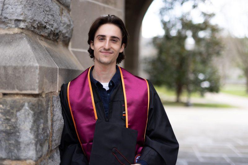 Wearing his graduation robe and stole, Jack smiles and leans against a Hokie Stone wall.